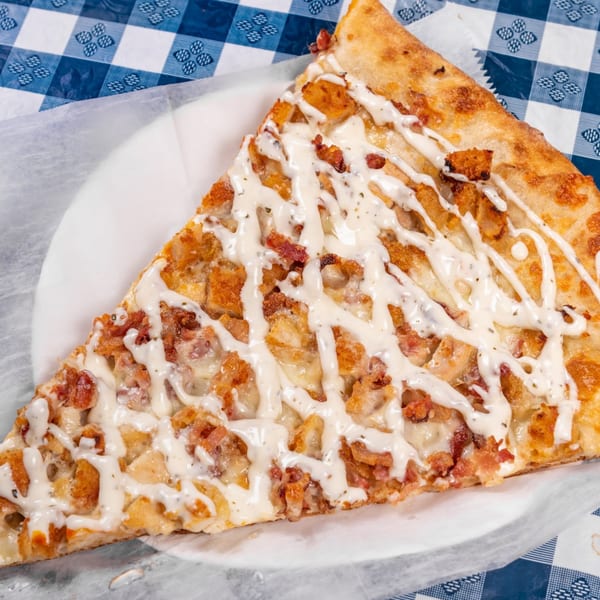 A slice of pizza with chicken, bacon, and a creamy white sauce on a blue and white checkered tablecloth.