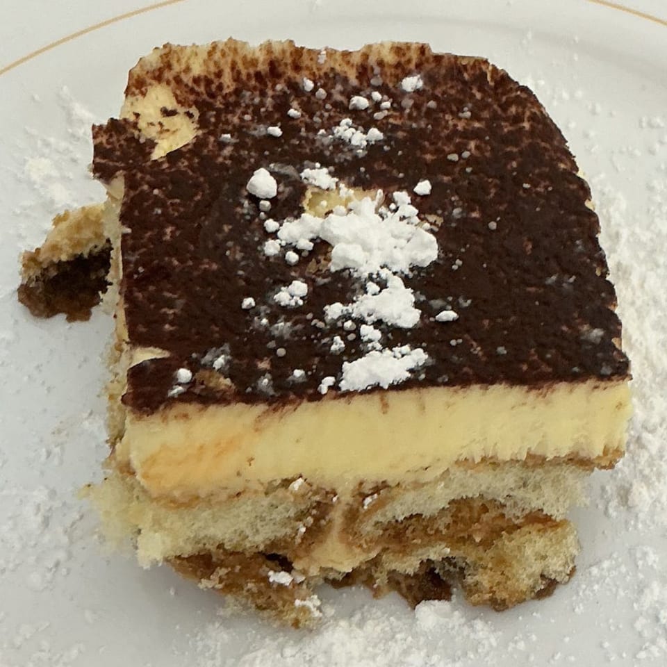 A slice of tiramisu with cocoa powder and powdered sugar on a white plate.