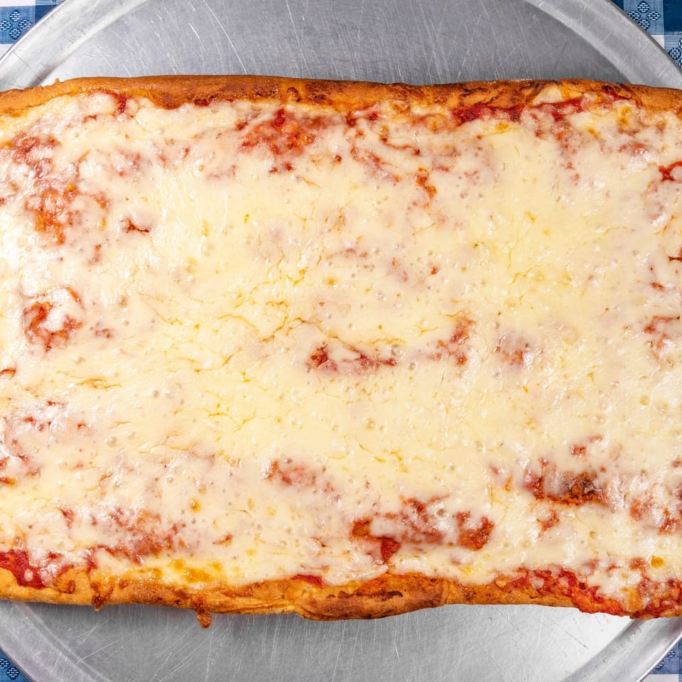 A rectangular cheese pizza with a golden crust sits on a metal tray, on a blue and white checkered tablecloth.